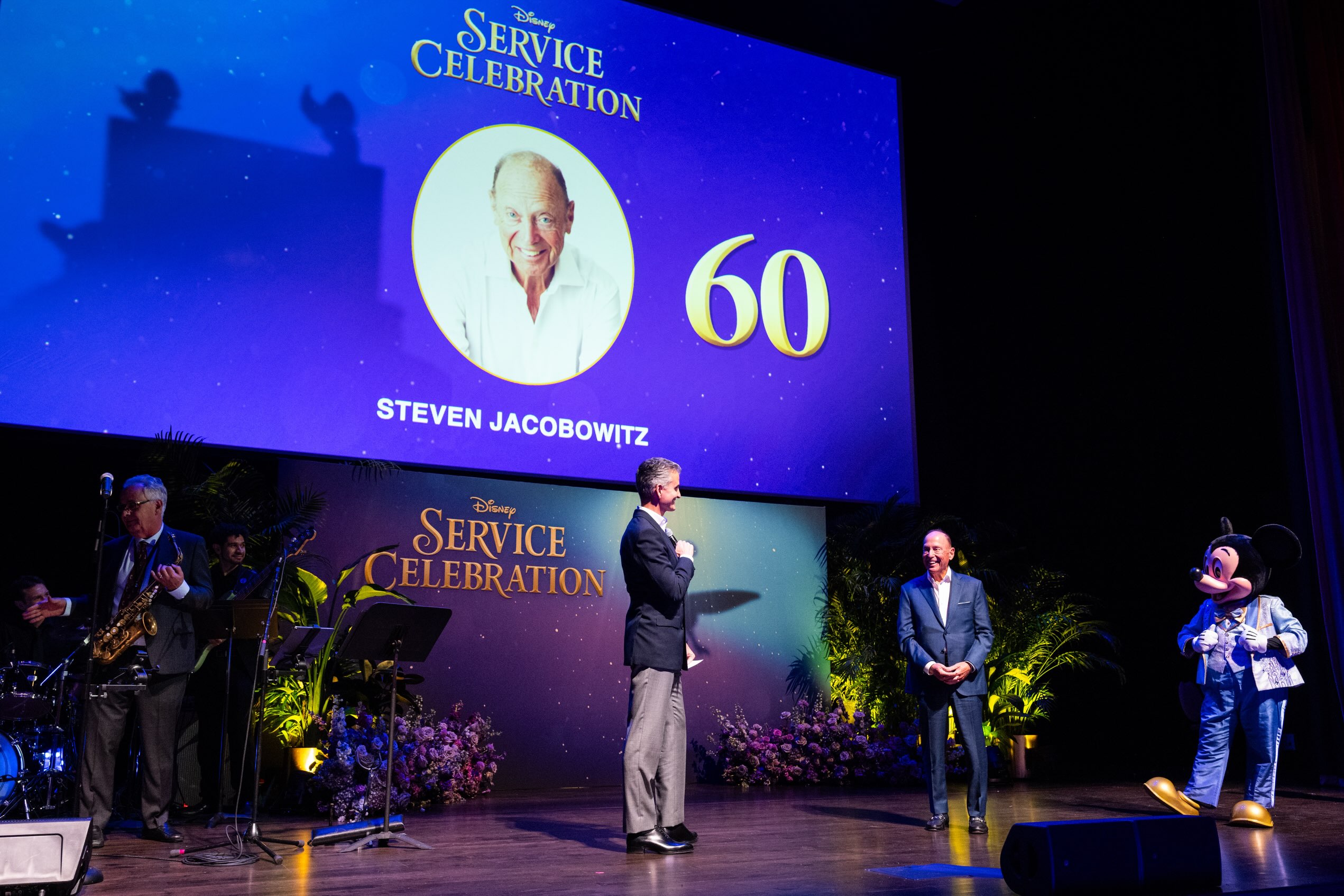 Josh D’Amaro speaks onstage during a Disney service celebration, with Mickey Mouse and an honoree nearby