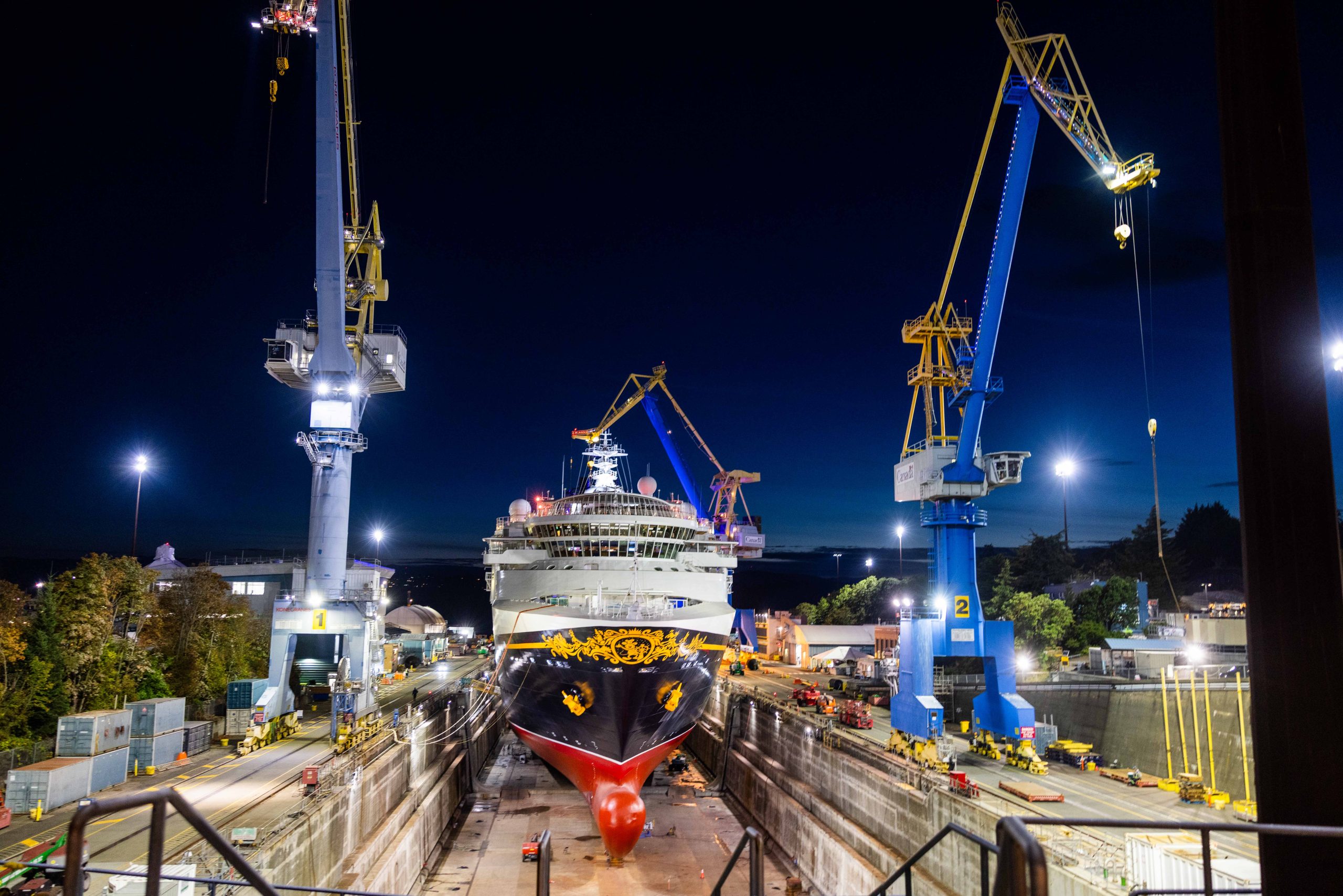 Disney Wonder sits in a dry dock at night between illuminated shipyard cranes