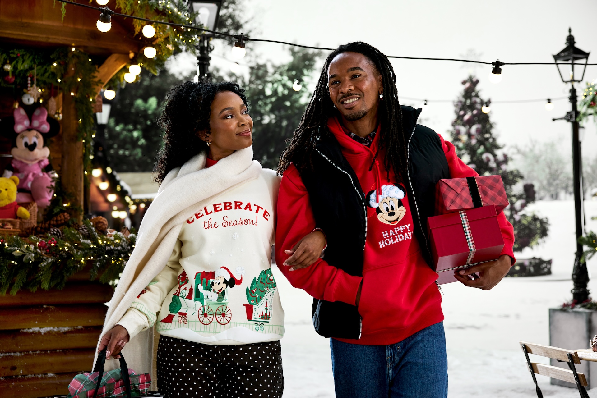 Two adults model Mickey and Minnie holiday apparel beside wrapped gifts in a decorated holiday setting