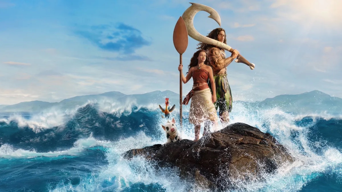 Live-action Moana stands with Maui, Heihei and Pua on rocks above the ocean