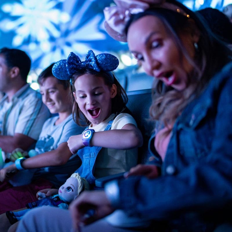 A child looks at a MagicBand on her wrist while seated with her family at Walt Disney World