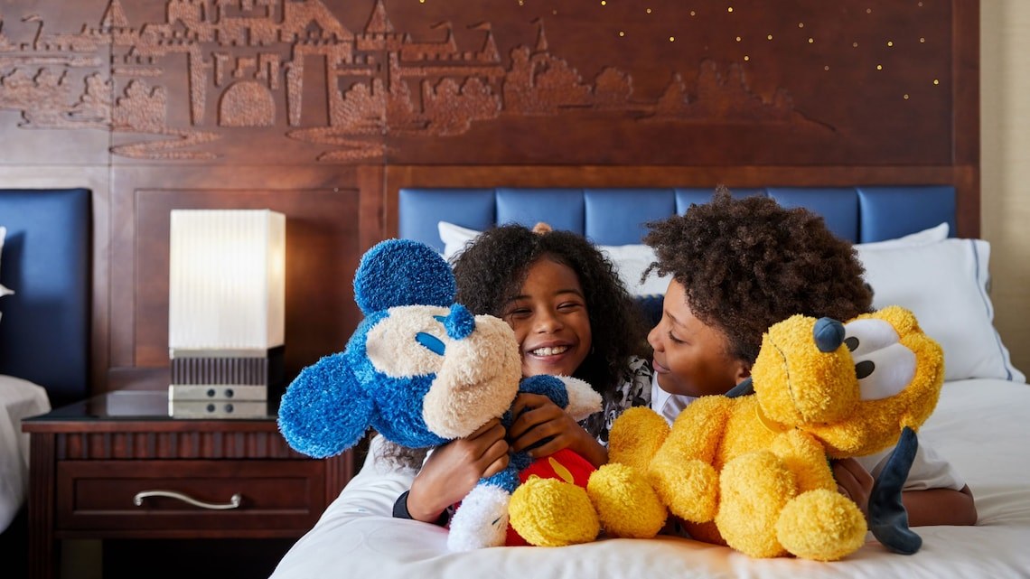 Two children hold Mickey Mouse and Pluto plush toys in a Disneyland Resort hotel room