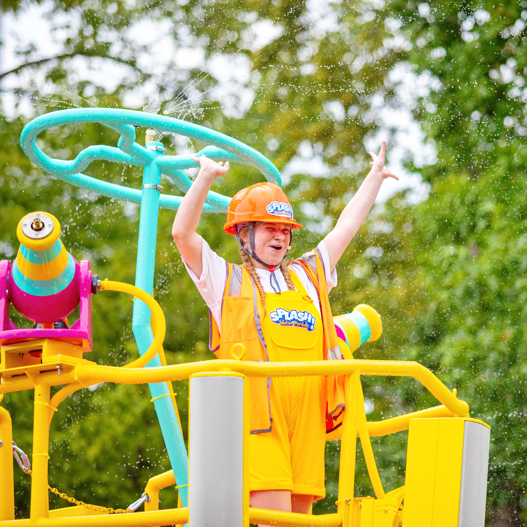 A SPLASH! Water Parade performer raises both arms as water sprays around a float