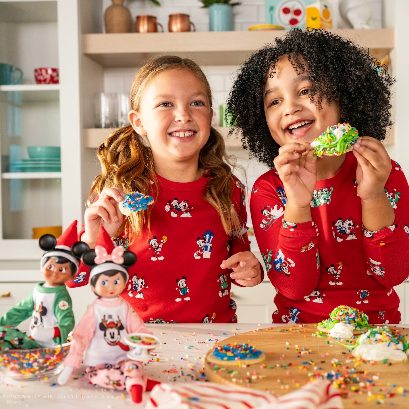 Children decorate cookies beside Elf on the Shelf dolls in Mickey and Minnie pajamas