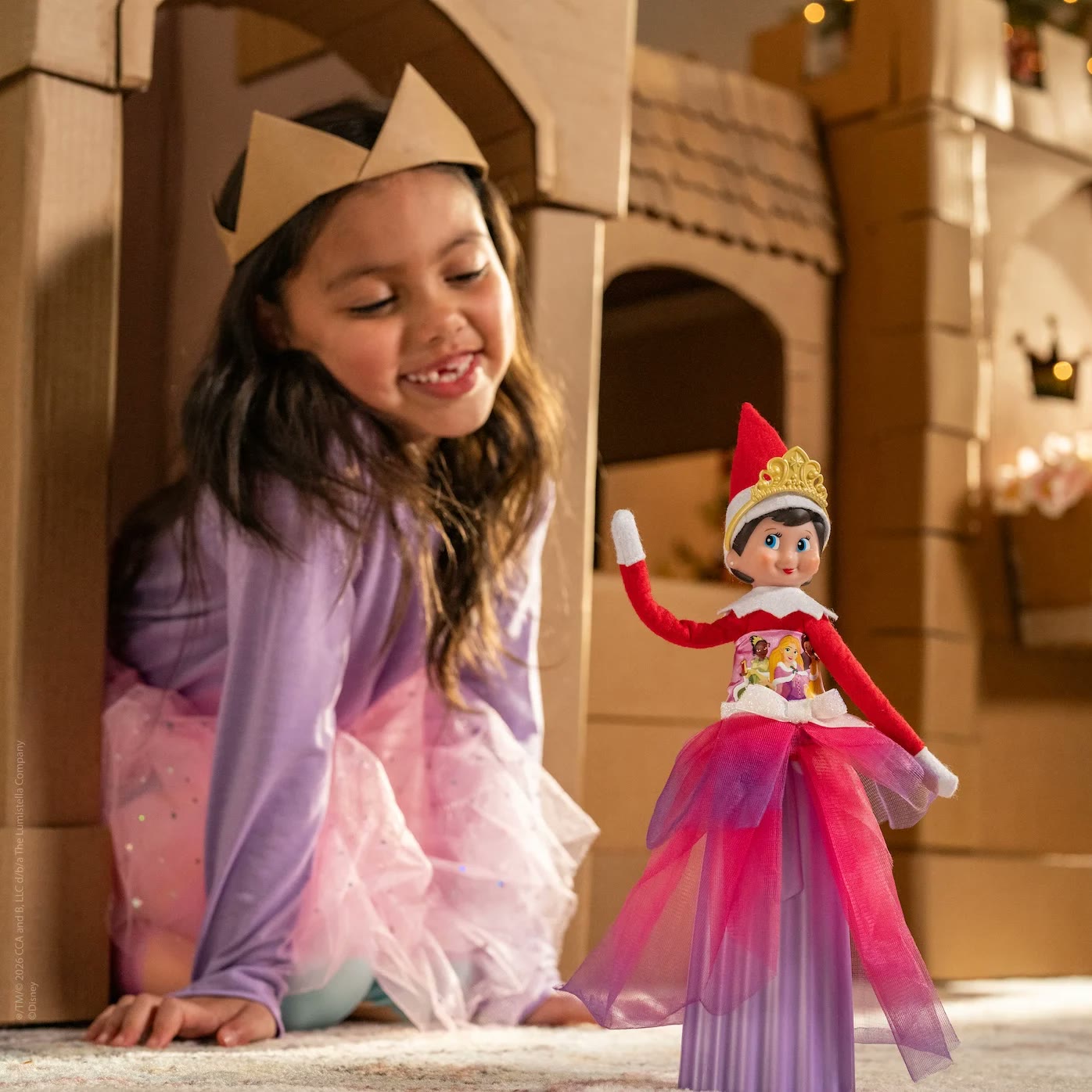 A child wears a paper crown beside an Elf on the Shelf in a Disney Princess outfit