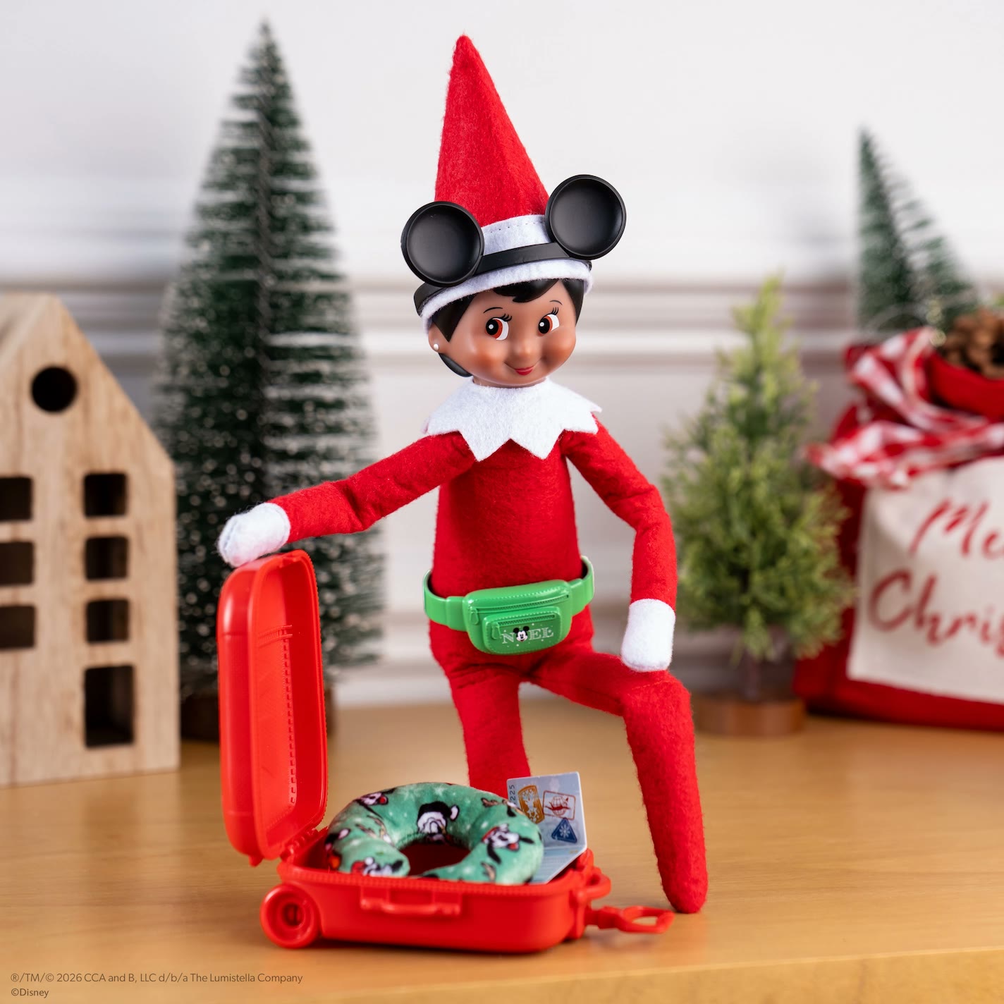 Elf on the Shelf wears Mickey Mouse ears beside a red suitcase