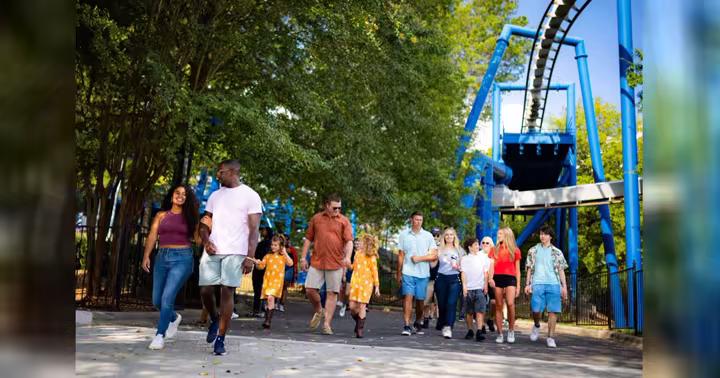 Guests walk past a blue roller coaster at a Six Flags park