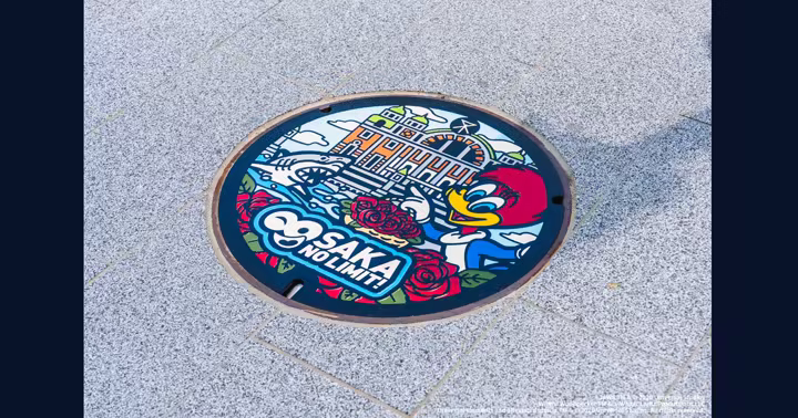 Universal Studios Japan anniversary manhole cover featuring Osaka City Central Public Hall, roses, Woody Woodpecker, and Jaws