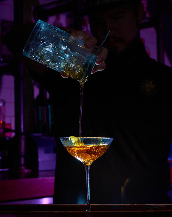An Edison bartender pours a cocktail into a coupe glass.
