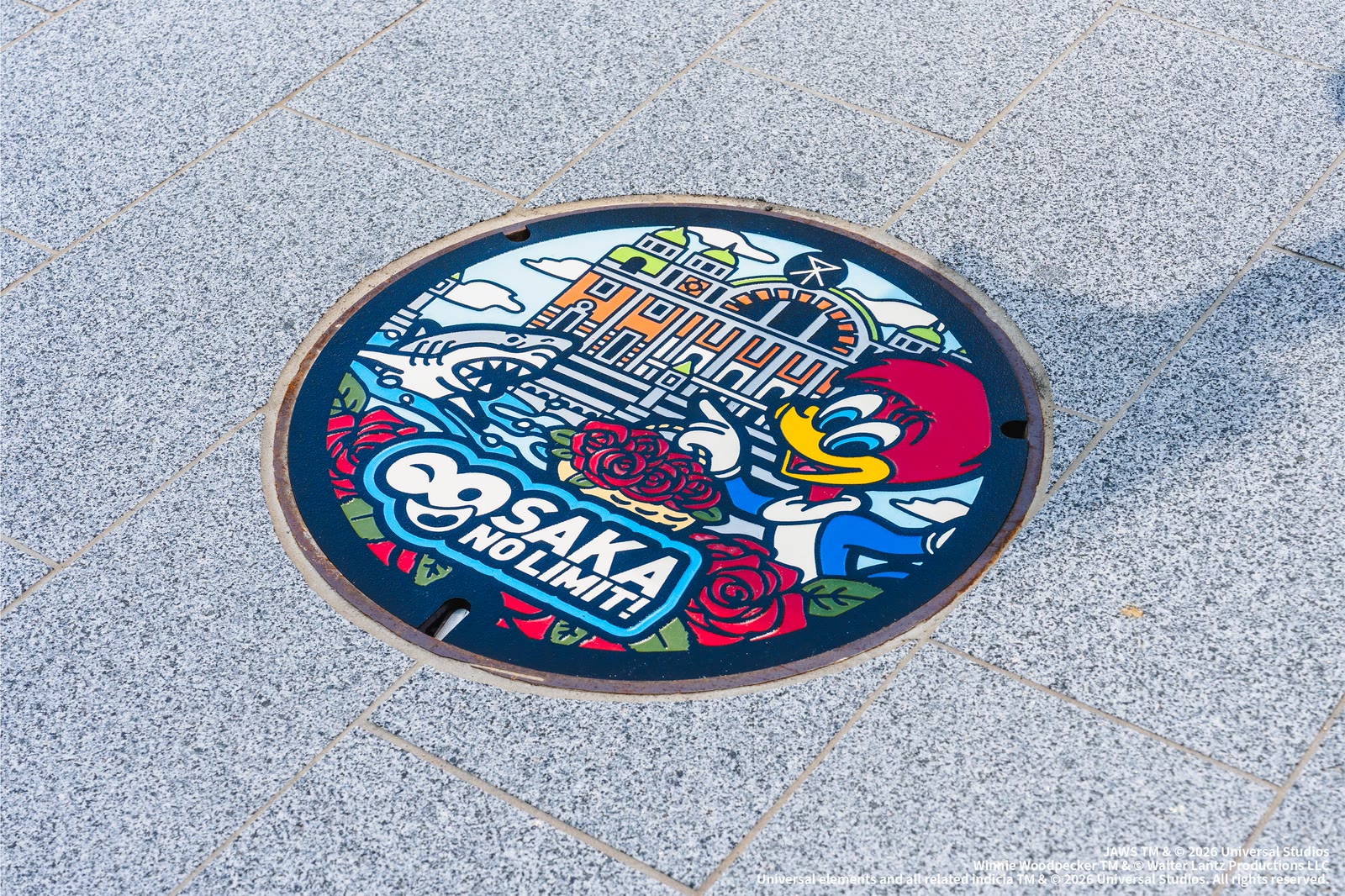 Universal Studios Japan anniversary manhole cover featuring Osaka City Central Public Hall, roses, Woody Woodpecker, and Jaws
