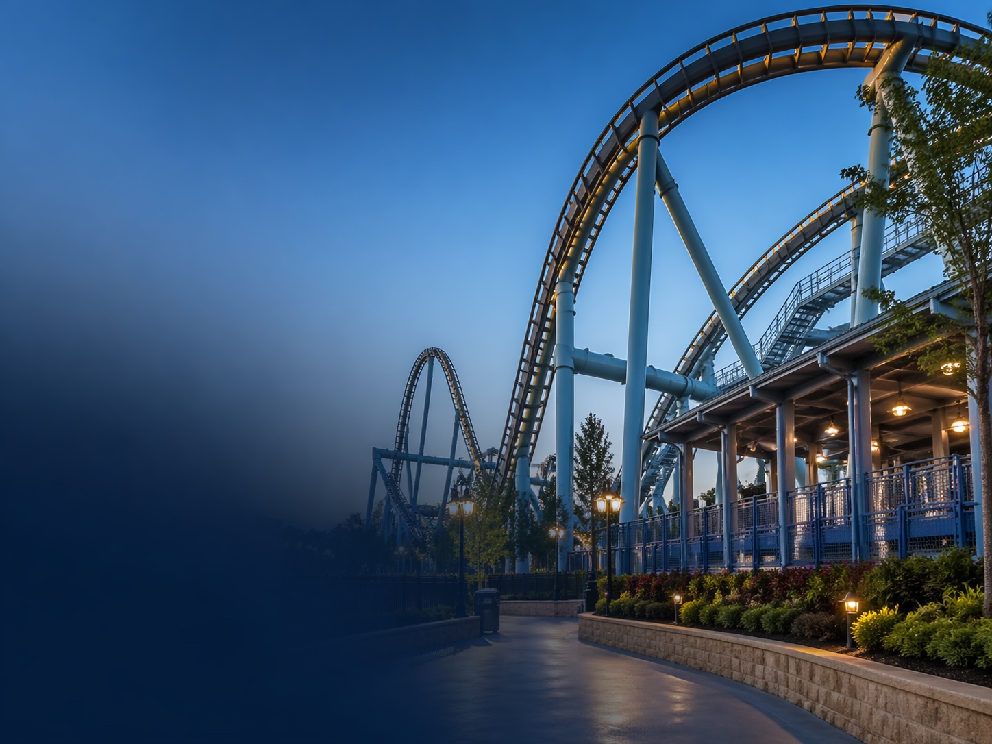 Large steel roller coaster track above an empty theme park pathway at twilight